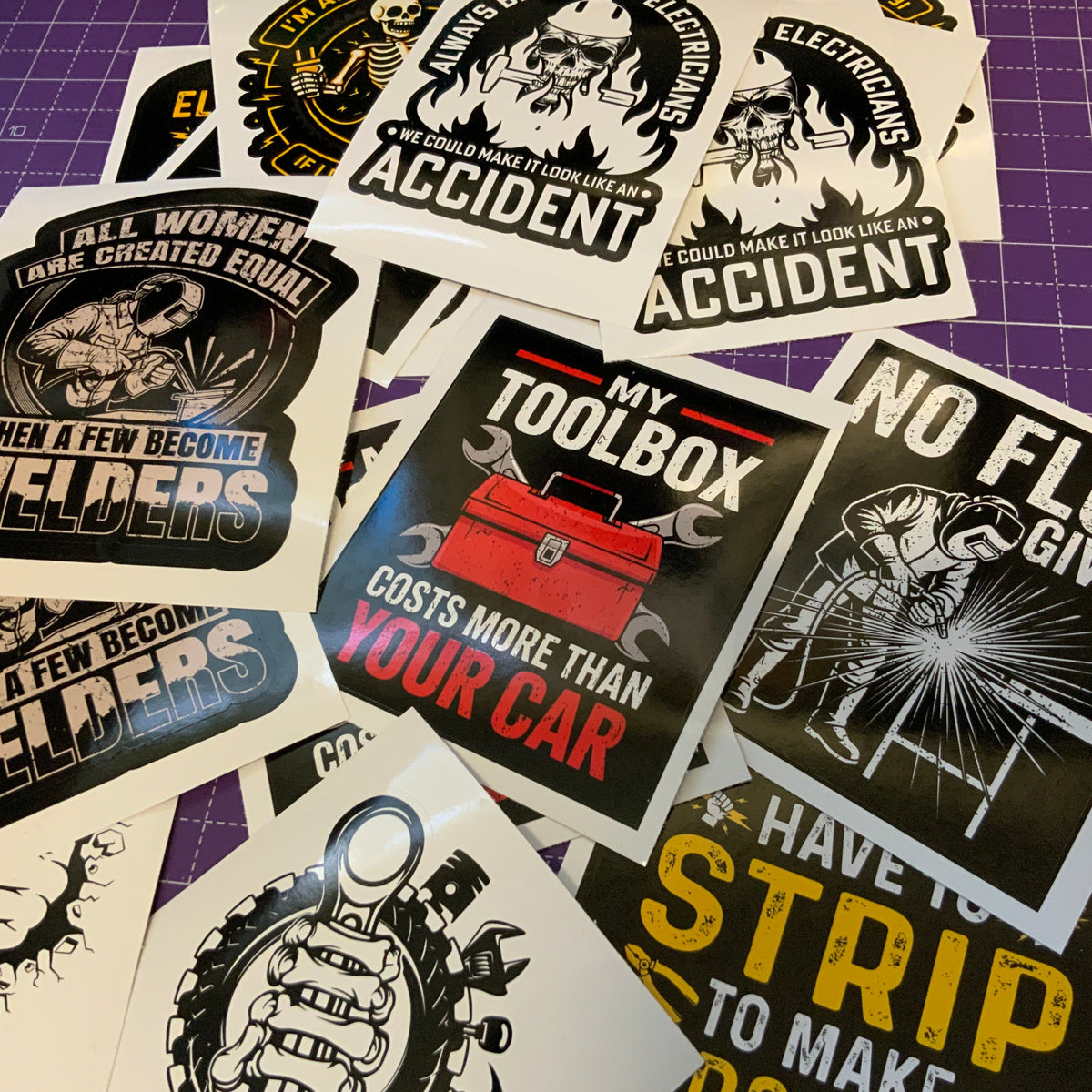 A collection of die-cut vinyl stickers featuring humorous and sarcastic automotive/welding-themed graphics, arranged on a purple cutting mat.