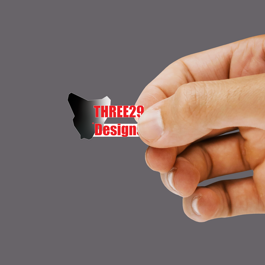 A close-up of a hand holding a small black die-cut sticker with the text 'THREE TWO Design' in red and white, set against a solid gray background.