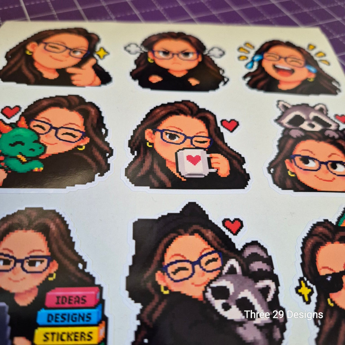 A sheet of pixel-art style stickers featuring a cartoon woman with glasses in multiple poses, including holding a green snake, a mug with a heart, and hugging a raccoon. The stickers are arranged on a white backing sheet, photographed on a purple cutting mat.