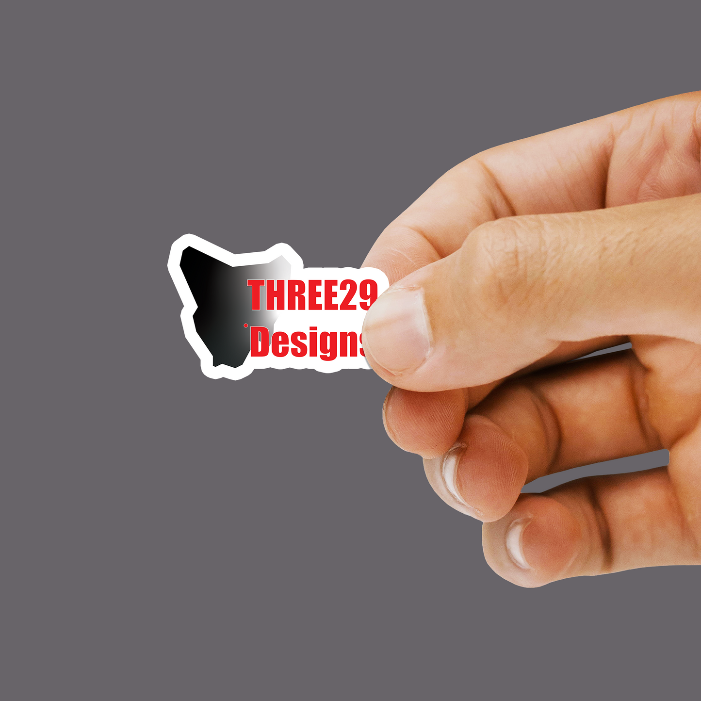 A hand holding a small die-cut sticker with a black-and-white silhouette shape and red text reading 'THREE29 Design' against a plain gray background.