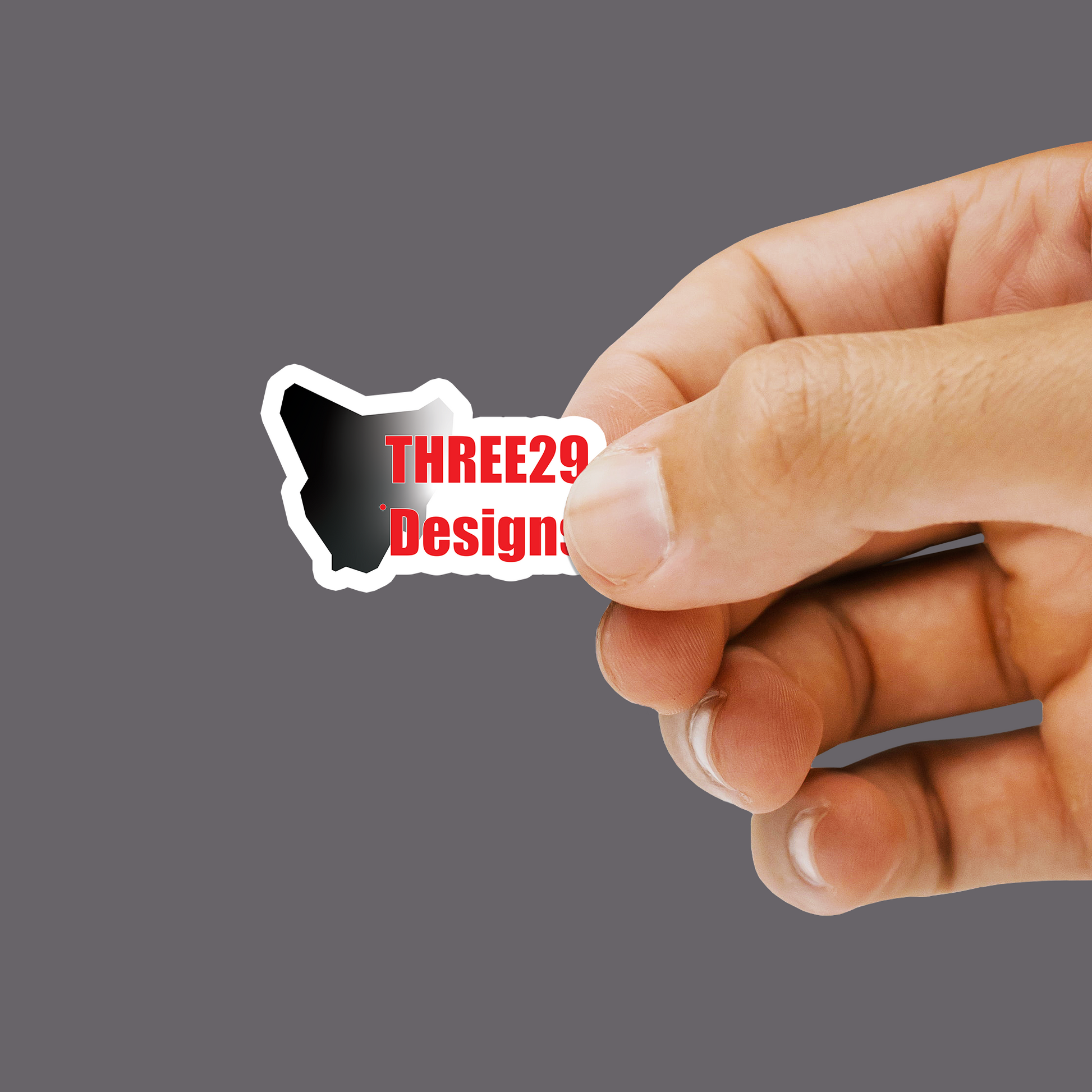 A hand holding a small die-cut sticker with a black-and-white silhouette shape and red text reading 'THREE29 Design' against a plain gray background.