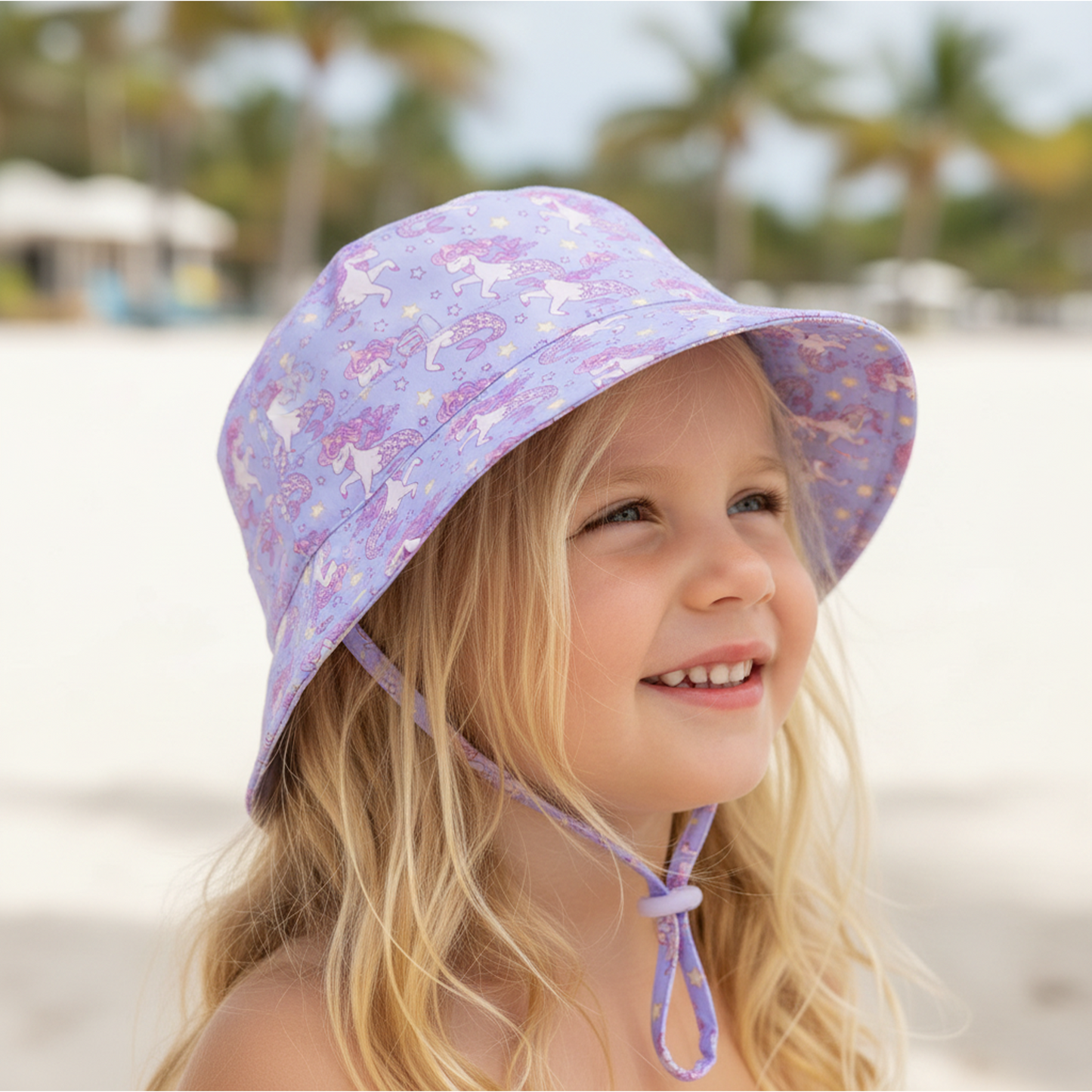 Young girl wearing a purple bucket hat with unicorn pattern on a beach.