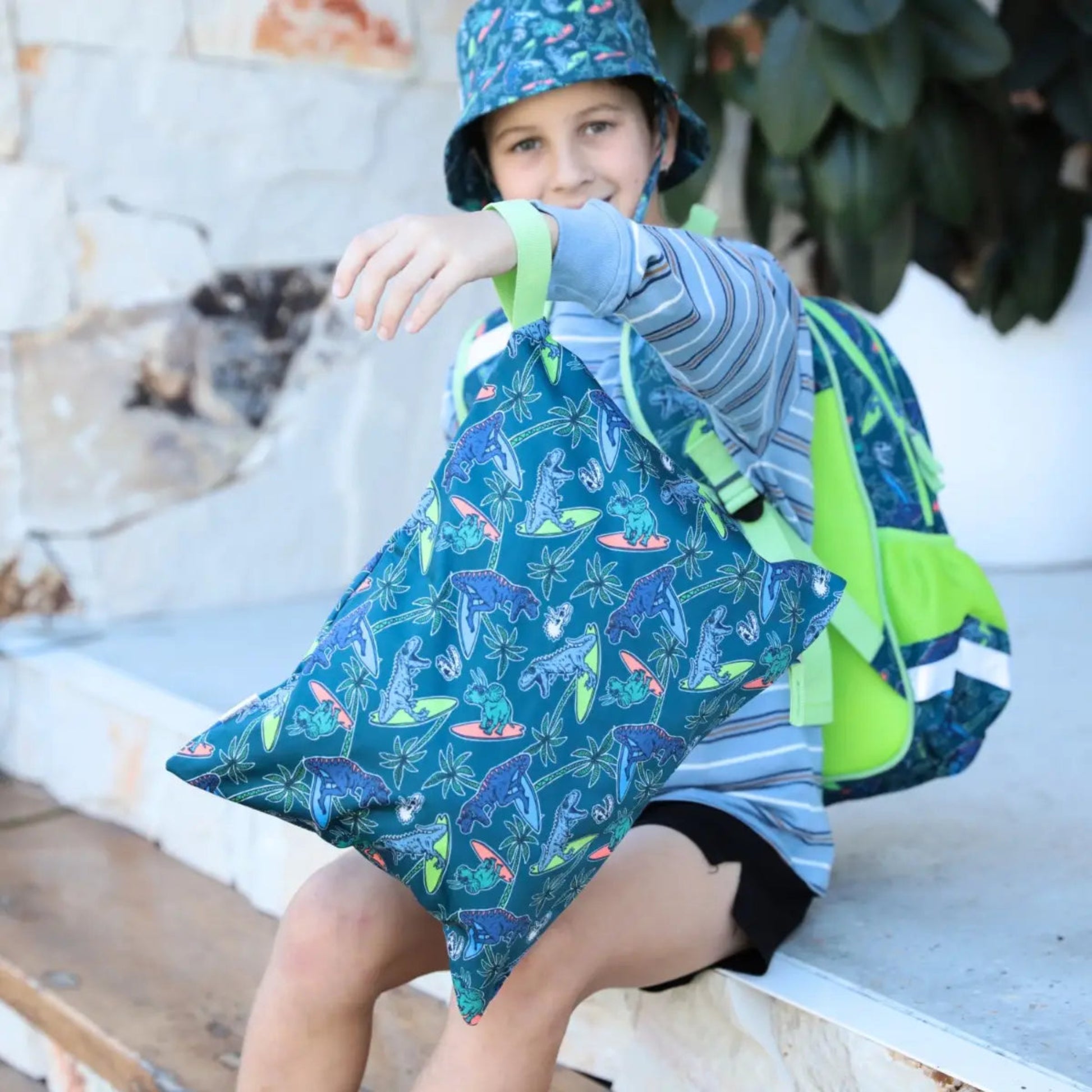 Child wearing a blue jacket with dinosaur pattern and green backpack sitting outdoors.